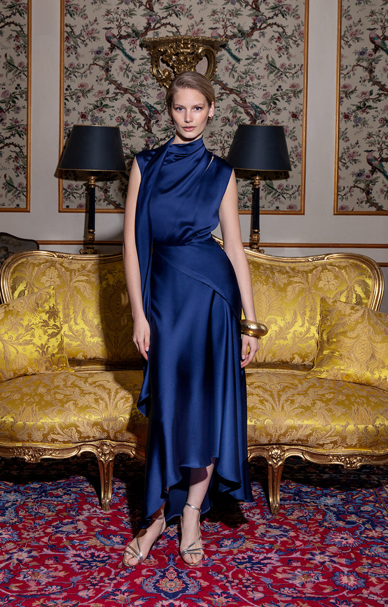 Woman in a blue dress standing in an ornate room with gold furniture and floral wallpaper.