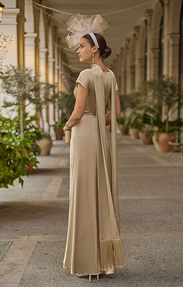 Woman in Couture Club 2G05 - Sand Satin Occasion Dress with Timeless Elegant Styling with a large white headpiece walking along a decorative path.