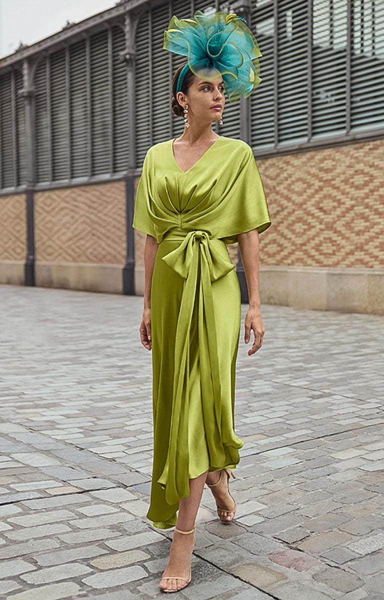 Woman in Couture Club 2G07 - Citrus Satin Occasion Dress with Contemporary Elegant Styling with a matching fascinator standing on a paved street.