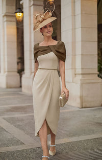 A woman wearing Rosa Clará Couture Club 2G12 - Sand and Chocolate Crepe Mermaid Midi Dress with Beaded Bodice &amp; Shawl, a matching straw fascinator hat, and holding a small metallic clutch, walking on a stone walkway.