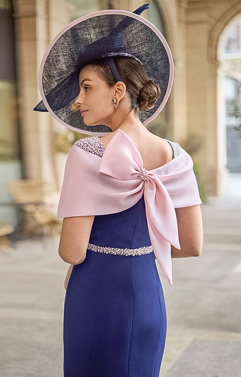 Photograph of the rear of a Woman wearing Couture Club 2G13 - Navy and Pale Rose Crepe Midi Dress with Elegant Colour-Block Design with feature pale rose / pink bow. She is standing in from of an elegant courtyard background  with potted olive trees.