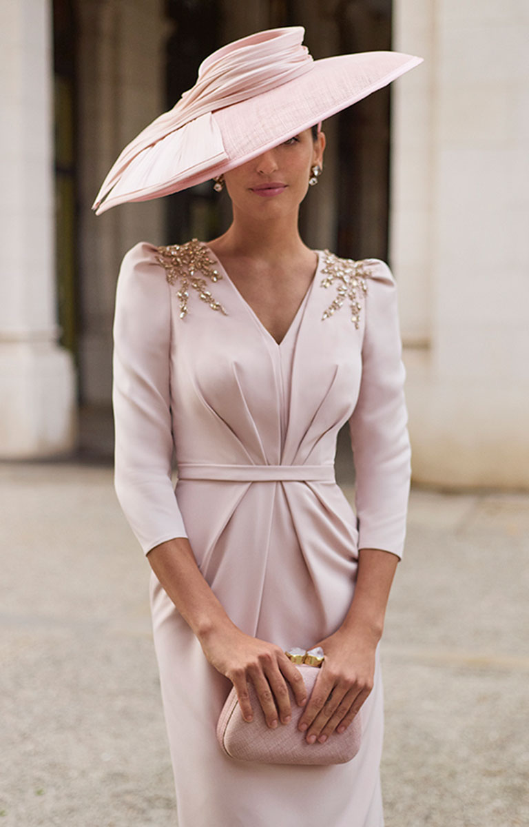 Woman in Couture Club 2G20 - Rosa Pastel Pink Crepe Occasion Dress with Timeless Feminine Styling with embellishments and a matching hat, holding a clutch, against a blurred architectural background.