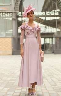 A woman stands outdoors wearing Rosa Clará Couture Club 2G25 - Crepe Satin Midi Dress with Floral Embellishment and Shawl with floral off-shoulder detailing, a matching pink fascinator hat, and holding a small pink clutch.