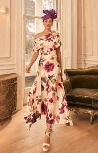 A woman models Veni Infantino 992827 - Champagne and Merlot Floral Satin Fit and Flare Midi Dress - a floral off-the-shoulder midi dress in a vintage-style room with a green velvet chaise lounge and wood floors. She is wearing a purple fascinator hat and white strappy heels.