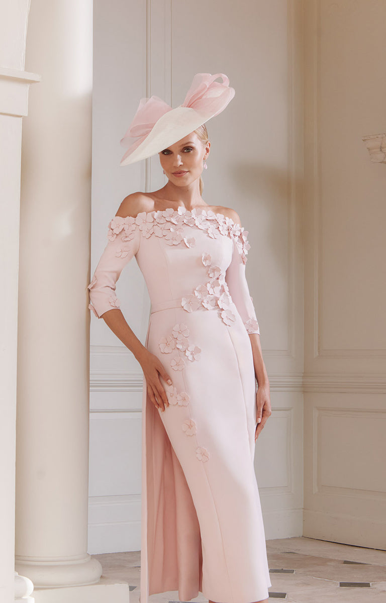 Woman in John Charles 29318 - Petal Pink Midi Dress with Off-the-Shoulder Neckline and 3D Floral Detail, standing against a white wall.