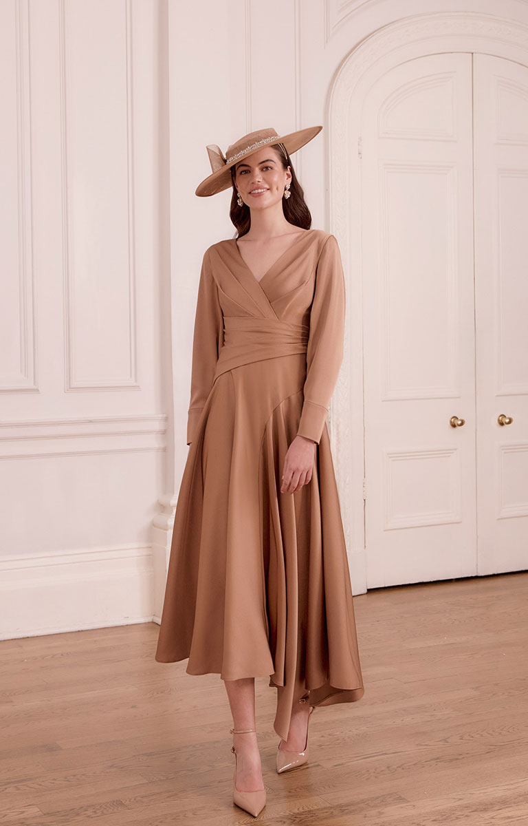 Woman in John Charles 29375 - Mocha Crepe Midi Dress with Pleated Waist and V-Neckline and hat standing in a room with white walls and wooden floor.