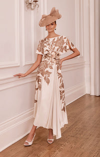 Woman in John Charles 66782B - Mocha &amp; Ecru Printed Draped Midi Dress with Floral Waist Detail and hat standing in an elegant room.