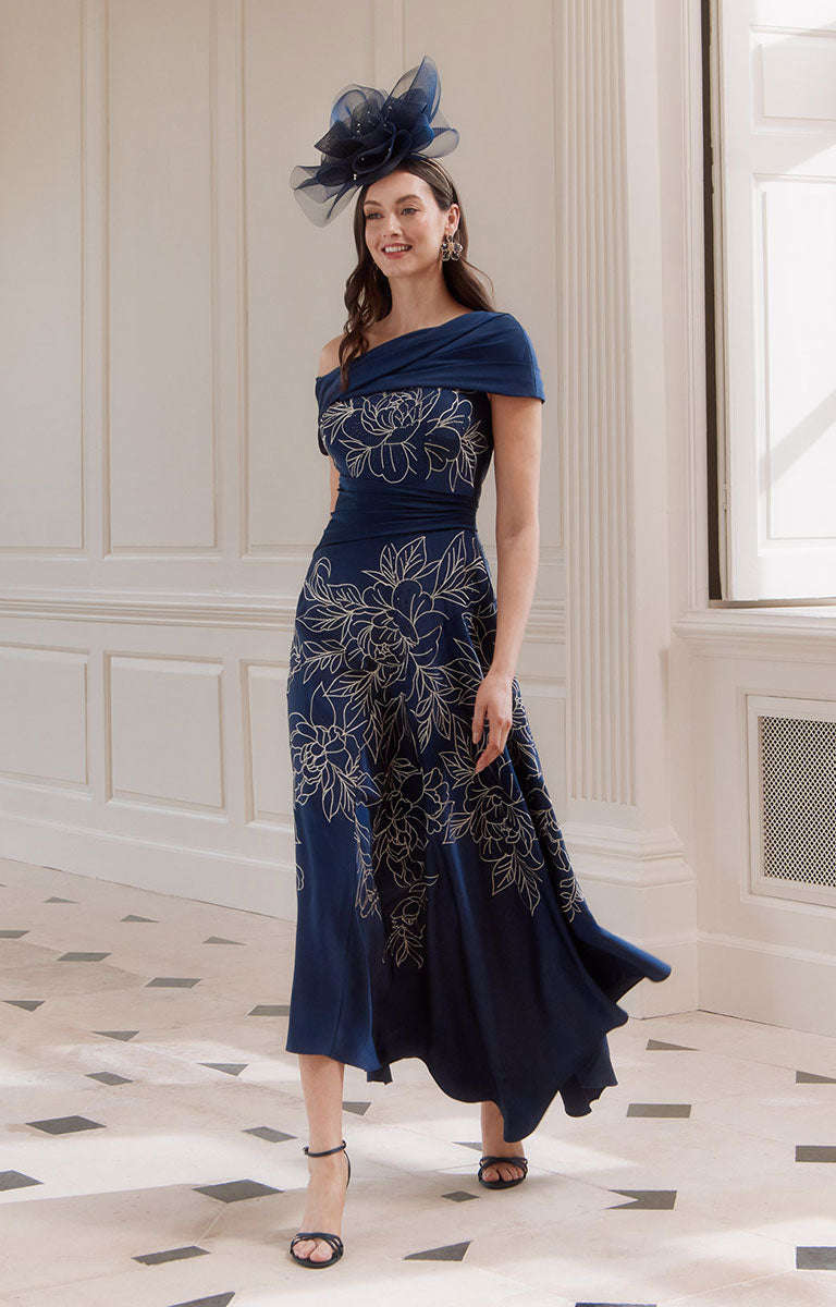 Woman in John Charles 66771 - Navy &amp; Oyster Midi Dress with Asymmetric Bardot Neckline and Floral Detail standing in an elegant room.
