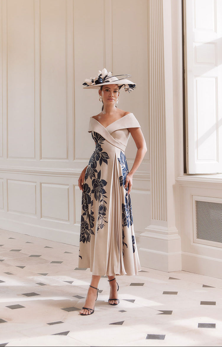 Woman wearing a John Charles dress called 66774 in the Navy / Oyster colourway. It is an elegant off-the-Shoulder Crepe Midi Dress with floral patterns in an elegant room. Perfect for the mother of the bride or groom at weddings or for ladies at formal events.