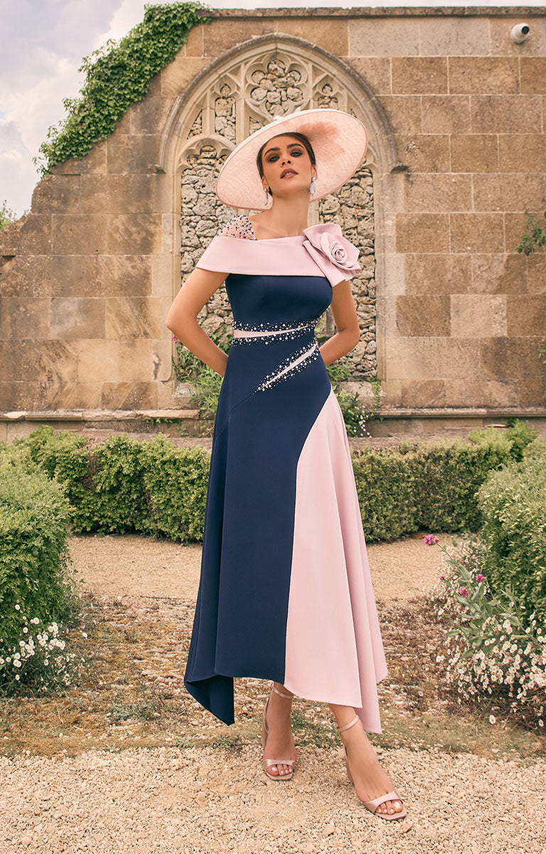 Woman in a Veni Infantino 992783 S – Navy / Blush Colour-Block Midi Dress with Draped Shoulder; Floral Motif wide hat standing in front of an old stone building.