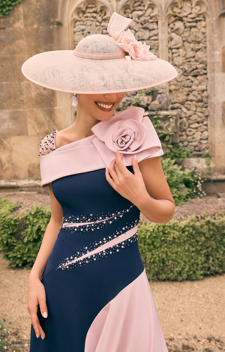 Woman in a Veni Infantino 992783 S – Navy / Blush Colour-Block Midi Dress with Draped Shoulder; Floral Motif wide hat standing in front of an old stone building.